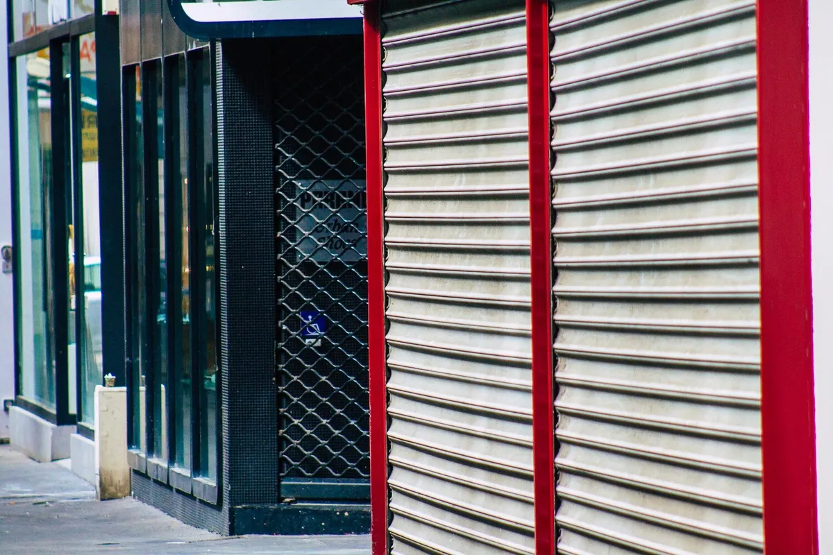 Rideau métallique à lames pleines encadré de rouge à côté d'une grille à mailles articulées de devanture