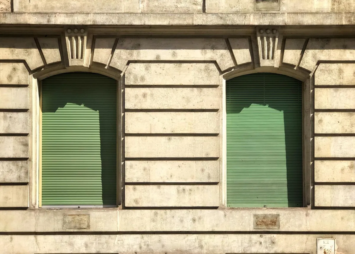 Deux volets roulants verts baissés dans des fenêtres cintrées d'une façade en pierre de taille à claveaux